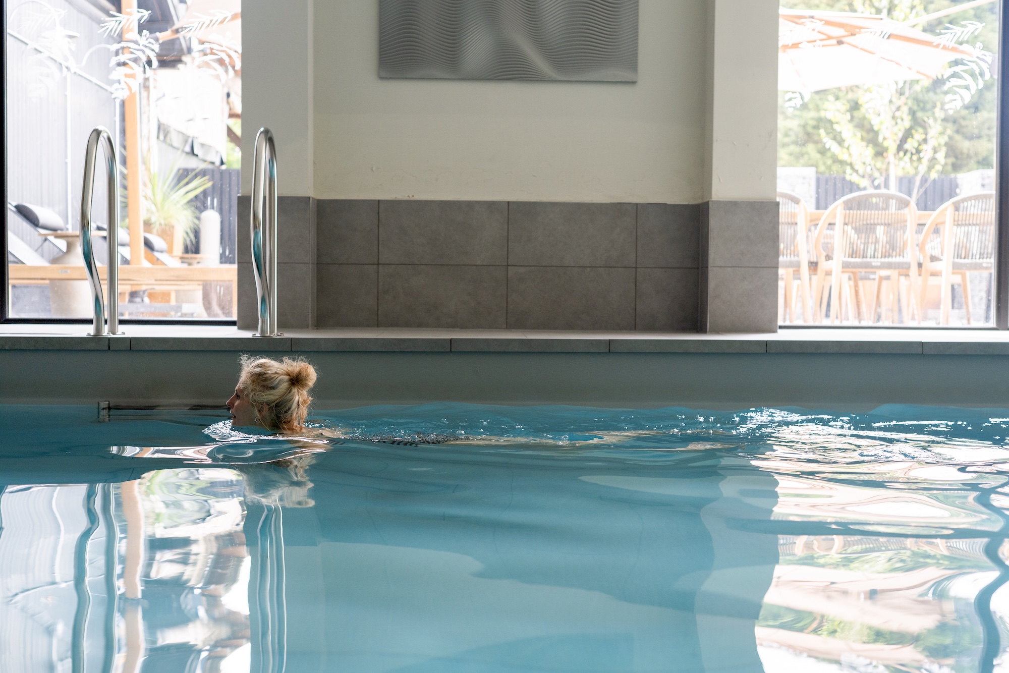 Swimmer in an indoor pool near the edge, with large windows and outdoor patio furniture visible outside.
