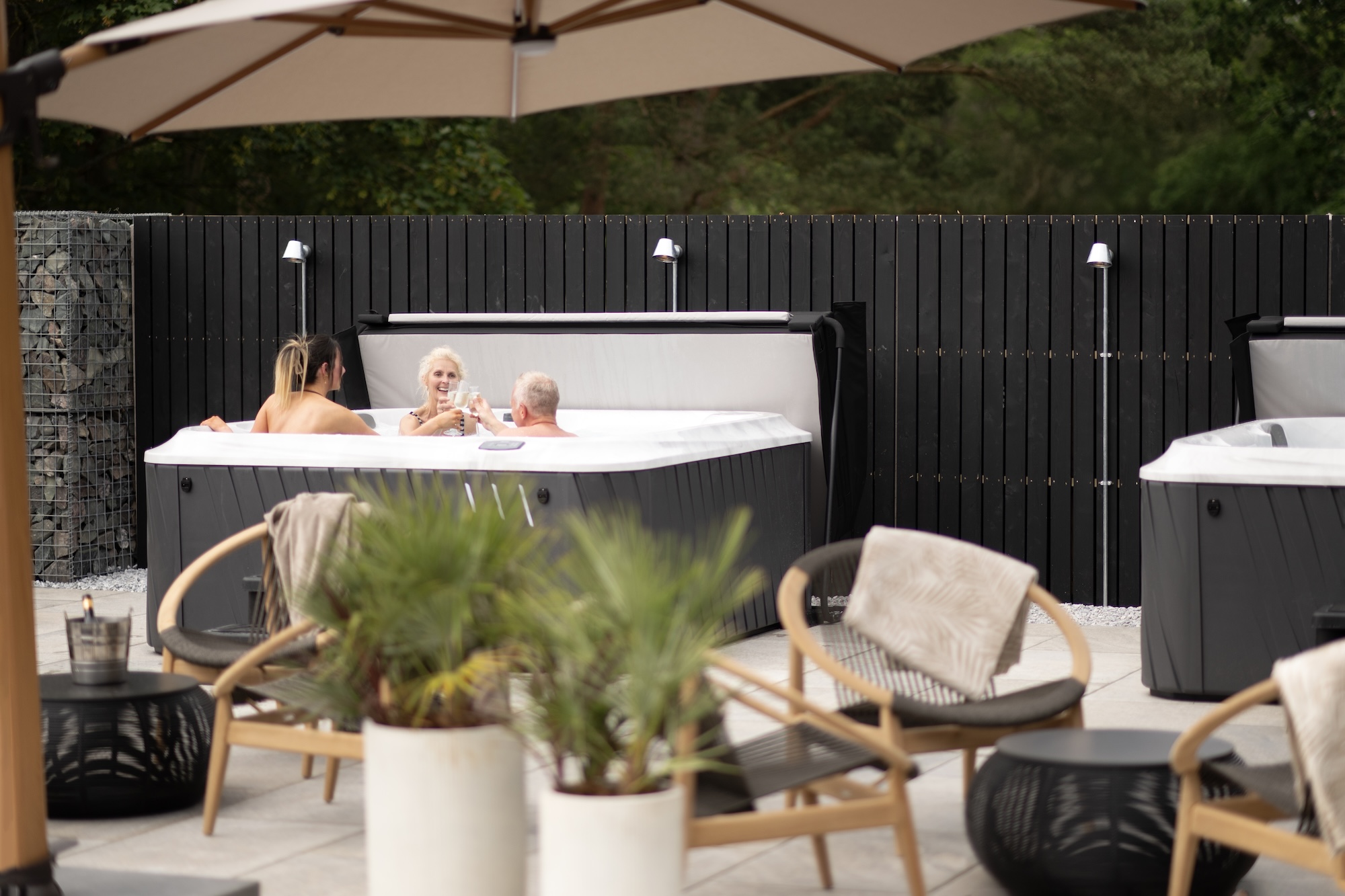 Three adults soak in an outdoor hot tub, clinking glasses beside a dark wooden fence and patio chairs.