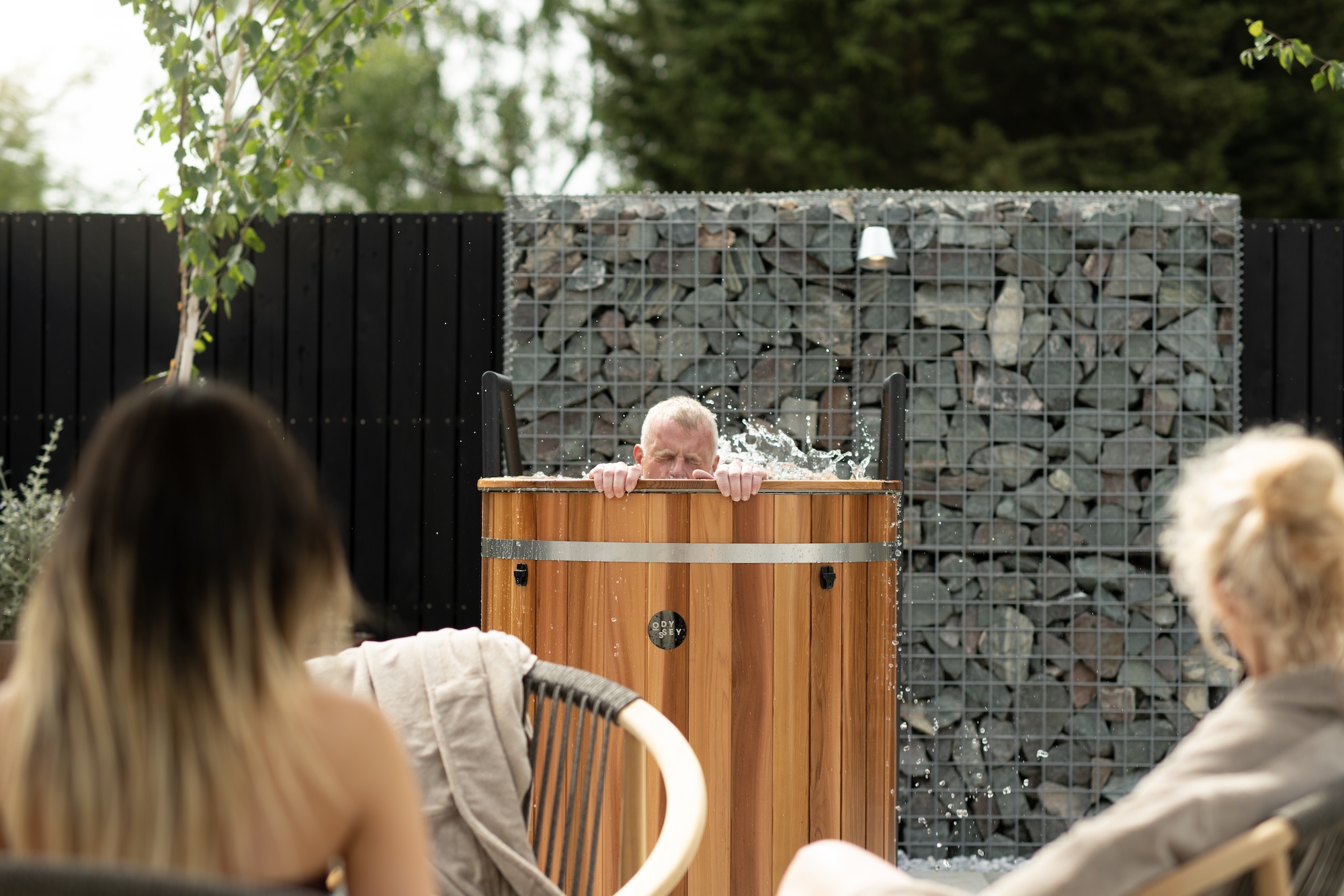 Man in a wooden outdoor hot tub splashes water, gripping the edge while onlookers watch from the foreground.