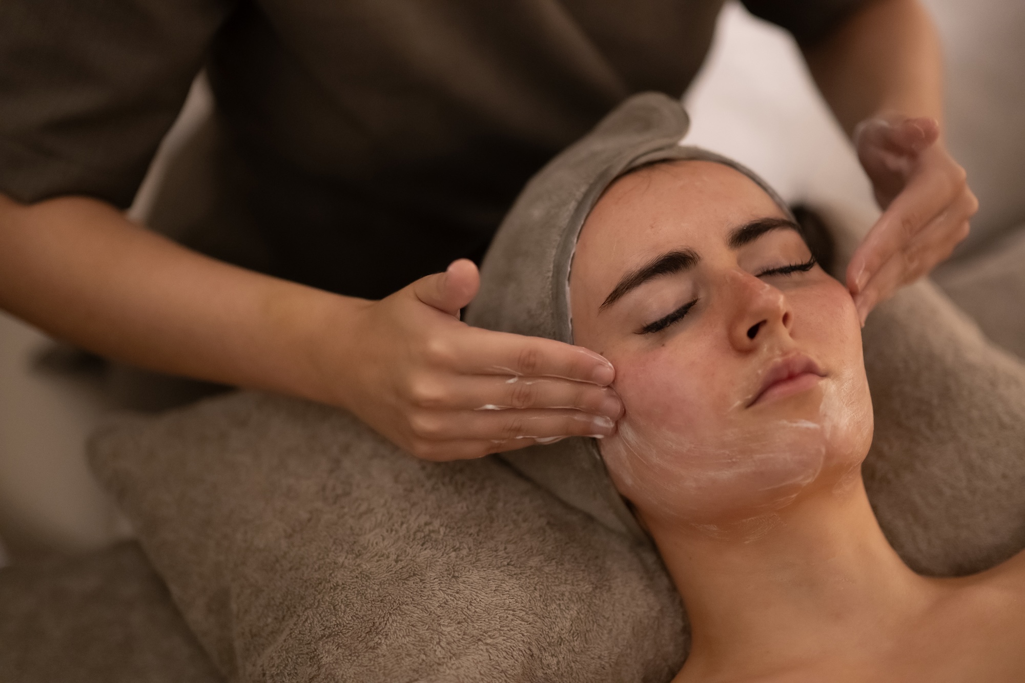 Person receiving a facial treatment: a therapist massages the client's head and neck while the client rests with eyes closed, a towel wrapped around the head and a facial mask applied at the cheeks and chin.