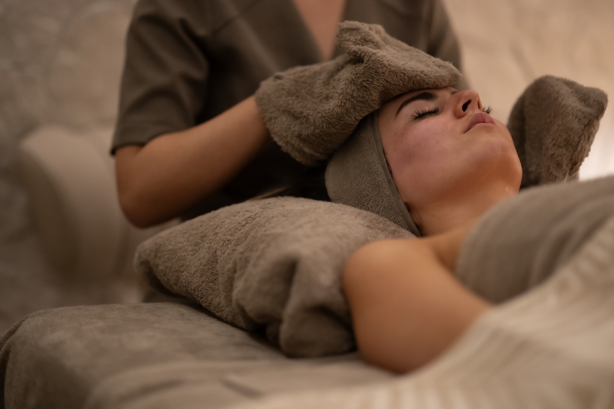 Person lying on a spa bed with a towel wrapped around their head, eyes closed during a facial treatment.
