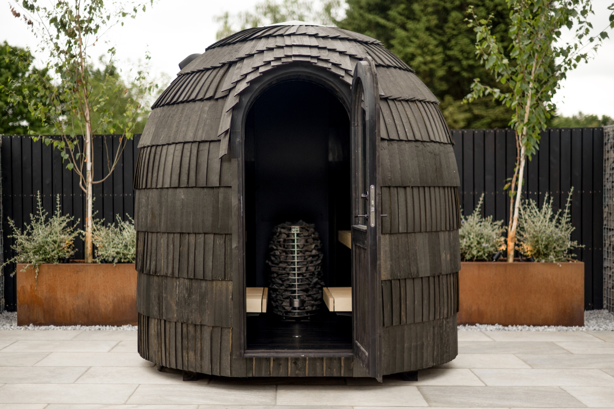 Round, domed wooden garden hut with an open door; interior benches and stacked firewood visible inside.