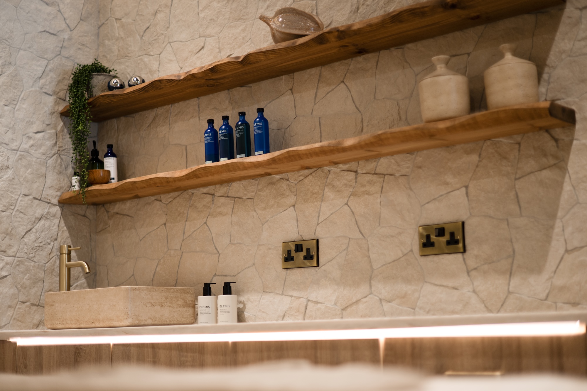 Stone-tiled bathroom wall with wooden floating shelves holding blue Elemis bottles and beige ceramic jars